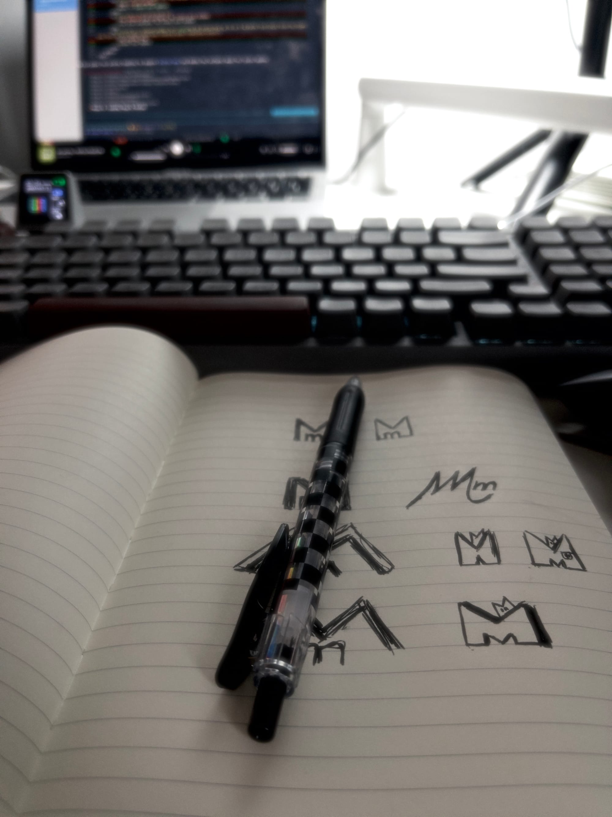 Photo of desk with laptop in the background and notebook, pen and keyboard.